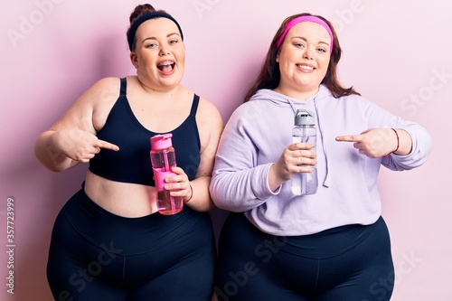 Young plus size twins wearing sportswear drinking bottle of water smiling happy pointing with hand and finger