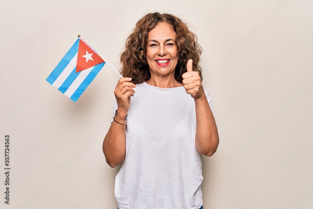 Middle age beautiful tourist woman holding cuban flag over isolated white background smiling happy and positive, thumb up doing excellent and approval sign Middle age beautiful tourist woman holding cuban flag over isolated white background smiling happy and positive, thumb up doing excellent and approval sign