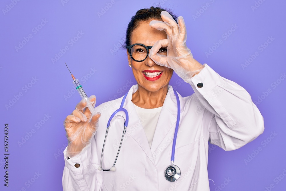 Middle age senior professional doctor woman holding syringe with medical vaccine with happy face smiling doing ok sign with hand on eye looking through fingers Middle age senior professional doctor woman holding syringe with medical vaccine with happy face smiling doing ok sign with hand on eye looking through fingers