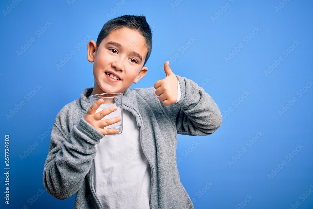 Adorable toddler smiling happy and confident. Standing with smile on face holding glass of healthy water to refreshment with thumb up doing ok sign over isolated blue background Adorable toddler smiling happy and confident. Standing with smile on face holding glass of healthy water to refreshment with thumb up doing ok sign over isolated blue background