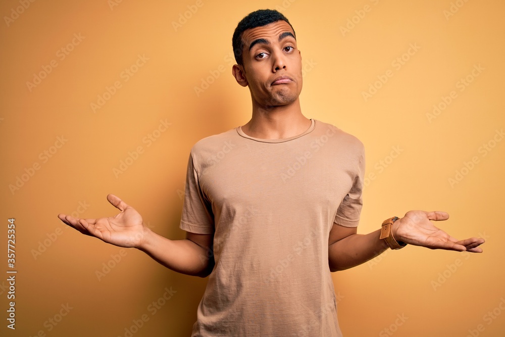 Young handsome african american man wearing casual t-shirt standing over yellow background clueless and confused with open arms, no idea concept. Young handsome african american man wearing casual t-shirt standing over yellow background clueless and confused with open arms, no idea concept.