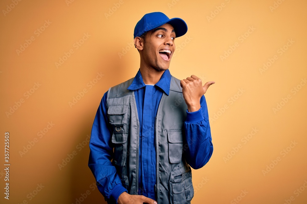 Young handsome african american handyman wearing worker uniform and cap smiling with happy face looking and pointing to the side with thumb up.