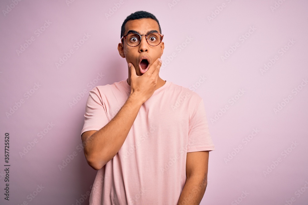 Handsome african american man wearing casual t-shirt and glasses over pink background Looking fascinated with disbelief, surprise and amazed expression with hands on chin Handsome african american man wearing casual t-shirt and glasses over pink background Looking fascinated with disbelief, surprise and amazed expression with hands on chin