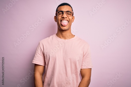 Handsome african american man wearing casual t-shirt and glasses over pink background sticking tongue out happy with funny expression. Emotion concept.