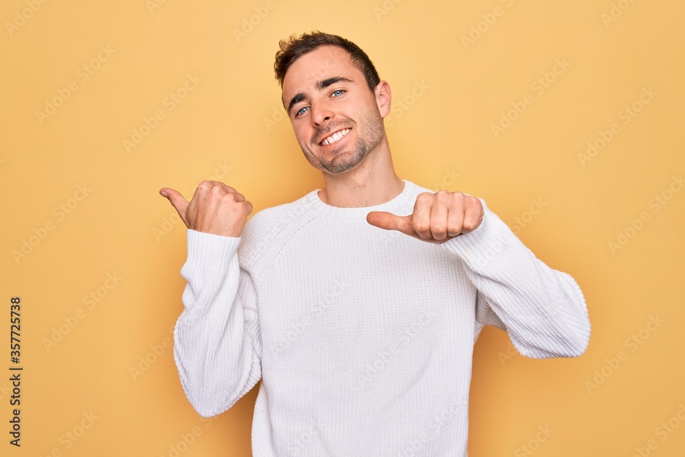 Young handsome man with blue eyes wearing casual sweater standing over yellow background Pointing to the back behind with hand and thumbs up, smiling confident