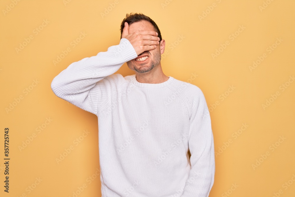 Young handsome man with blue eyes wearing casual sweater standing over yellow background smiling and laughing with hand on face covering eyes for surprise. Blind concept. Young handsome man with blue eyes wearing casual sweater standing over yellow background smiling and laughing with hand on face covering eyes for surprise. Blind concept.