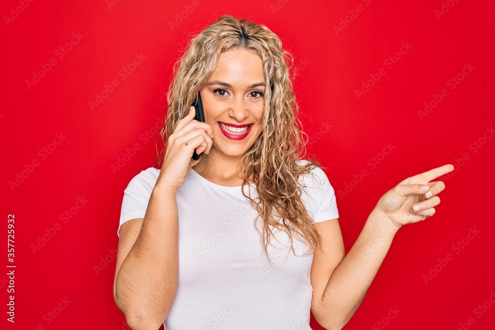 Beautiful blonde woman having conversation talking on the smartphone over red background Smiling happy pointing with hand and finger to the side Beautiful blonde woman having conversation talking on the smartphone over red background Smiling happy pointing with hand and finger to the side