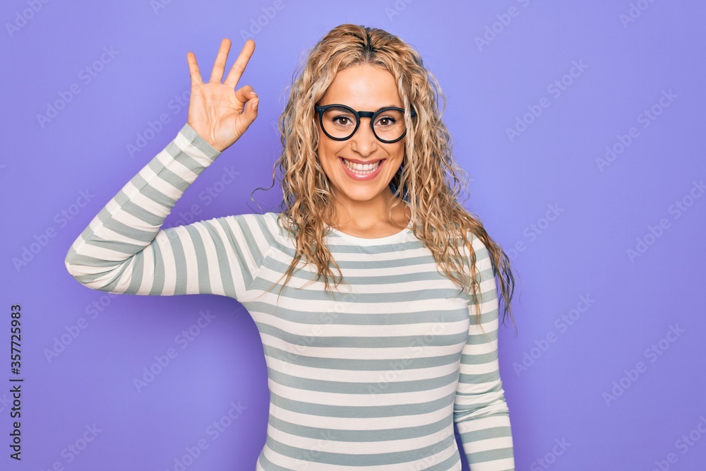 Beautiful blonde woman wearing casual striped t-shirt and glasses over purple background smiling positive doing ok sign with hand and fingers. Successful expression. Beautiful blonde woman wearing casual striped t-shirt and glasses over purple background smiling positive doing ok sign with hand and fingers. Successful expression.