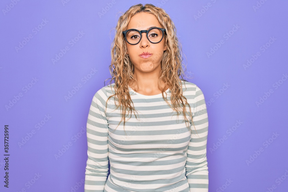 Beautiful blonde woman wearing casual striped t-shirt and glasses over purple background puffing cheeks with funny face. Mouth inflated with air, crazy expression. Beautiful blonde woman wearing casual striped t-shirt and glasses over purple background puffing cheeks with funny face. Mouth inflated with air, crazy expression.