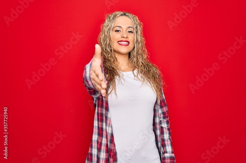 Young beautiful blonde woman wearing casual shirt standing over isolated red background smiling friendly offering handshake as greeting and welcoming. Successful business.