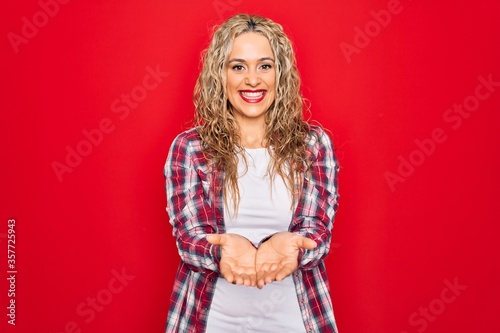 Young beautiful blonde woman wearing casual shirt standing over isolated red background Smiling with hands palms together receiving or giving gesture. Hold and protection