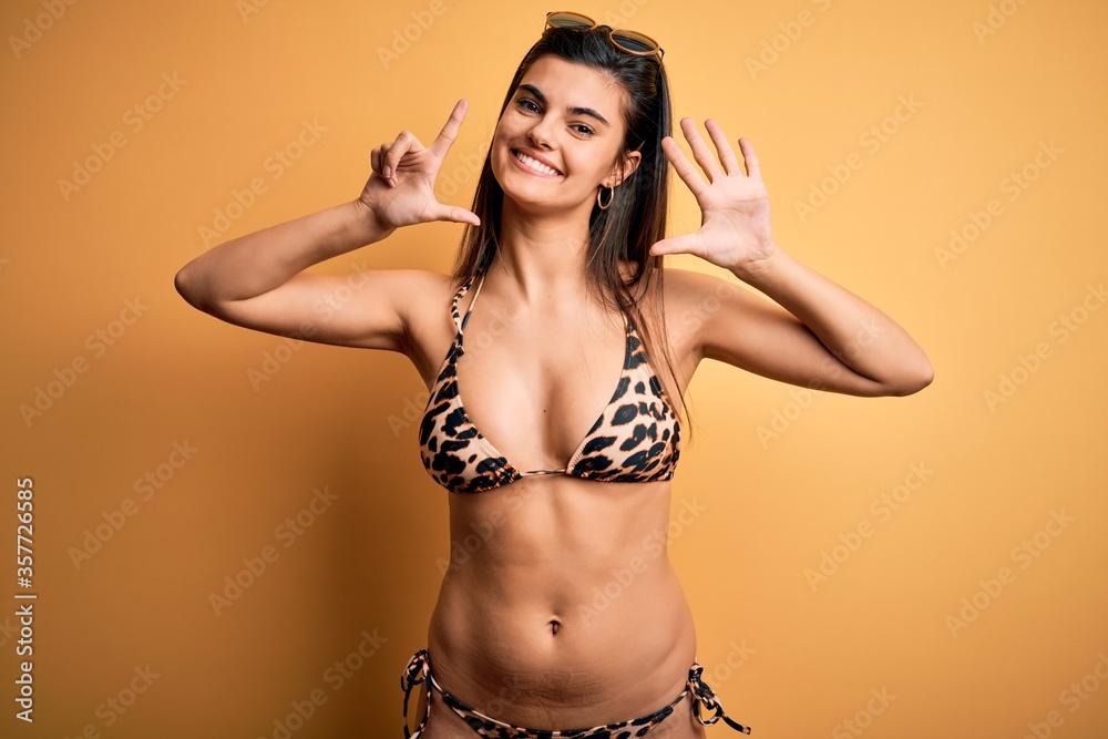 Φωτογραφία Young beautiful brunette woman on vacation wearing swimwear bikini over yellow background showing and pointing up with fingers number seven while smiling confident and happy