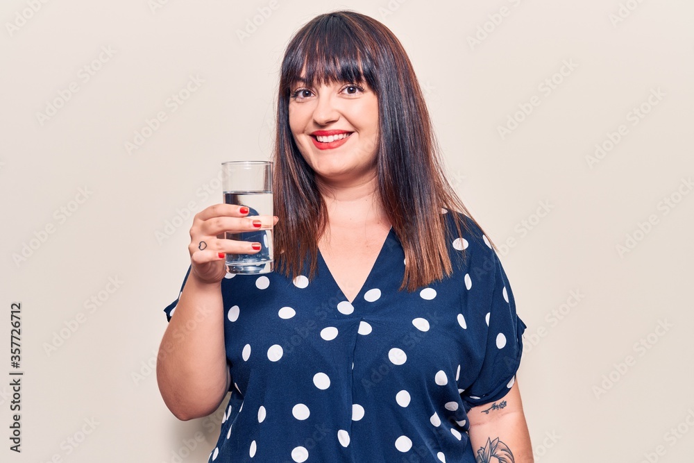 Young plus size woman drinking glass of water looking positive and happy standing and smiling with a confident smile showing teeth Young plus size woman drinking glass of water looking positive and happy standing and smiling with a confident smile showing teeth