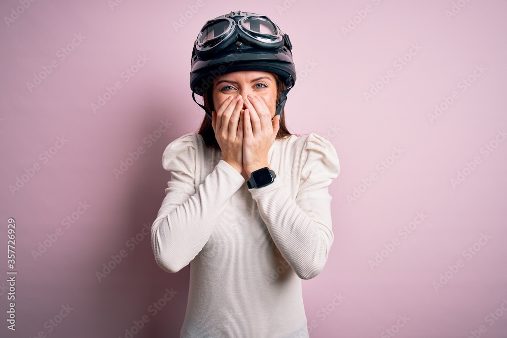 Young beautiful motorcyclist woman with blue eyes wearing moto helmet over pink background laughing and embarrassed giggle covering mouth with hands, gossip and scandal concept