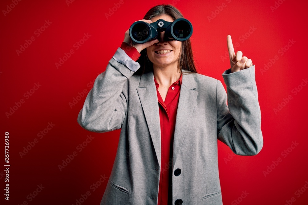 Young beautiful woman with blue eyes using binoculars over isolated red background surprised with an idea or question pointing finger with happy face, number one Young beautiful woman with blue eyes using binoculars over isolated red background surprised with an idea or question pointing finger with happy face, number one