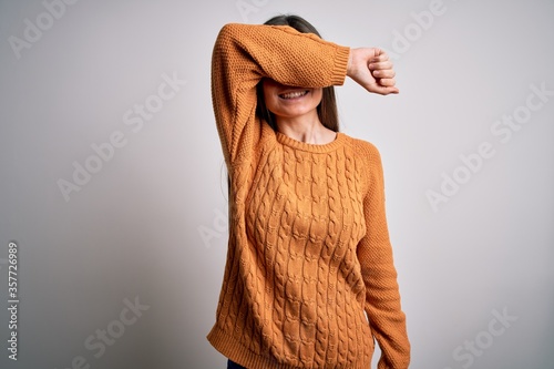 Young beautiful woman with blue eyes wearing casual sweater standing over white background covering eyes with arm smiling cheerful and funny. Blind concept.