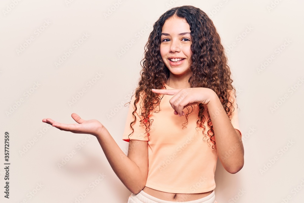 Beautiful kid girl with curly hair wearing casual clothes amazed and smiling to the camera while presenting with hand and pointing with finger.