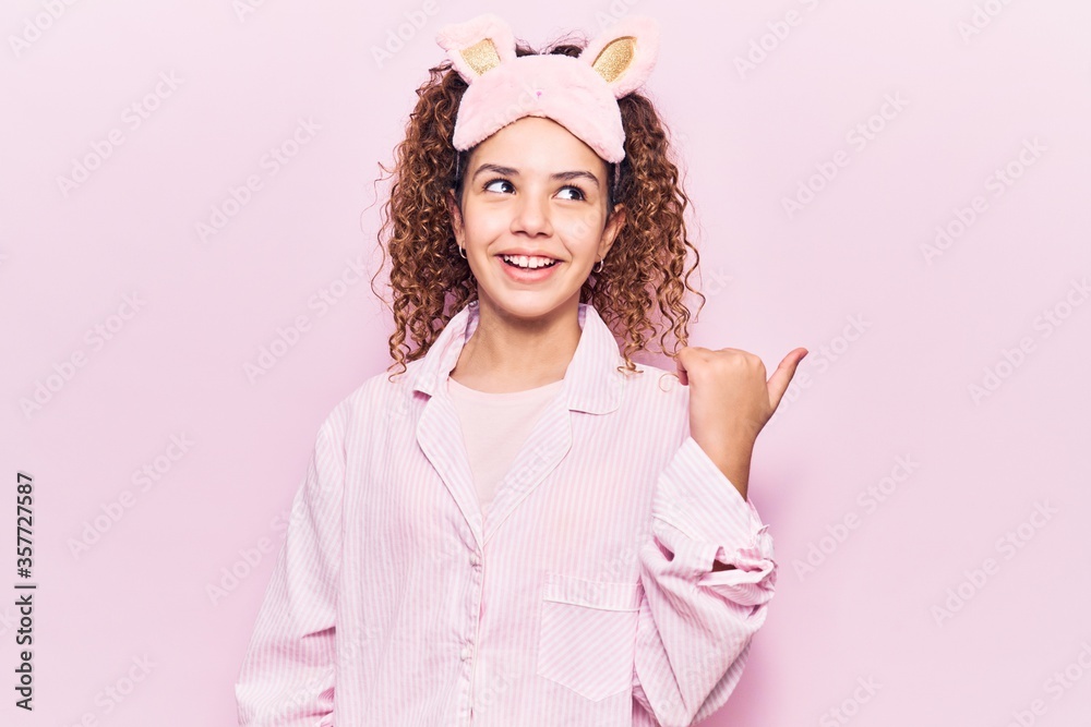 Beautiful kid girl with curly hair wearing sleep mask and pajamas smiling with happy face looking and pointing to the side with thumb up. Beautiful kid girl with curly hair wearing sleep mask and pajamas smiling with happy face looking and pointing to the side with thumb up.
