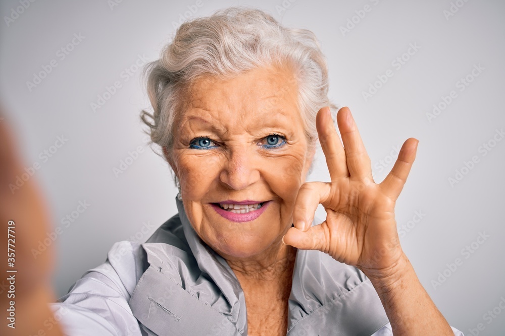 Senior beautiful grey-haired woman making selfie by camera over isolated white background doing ok sign with fingers, excellent symbol Senior beautiful grey-haired woman making selfie by camera over isolated white background doing ok sign with fingers, excellent symbol