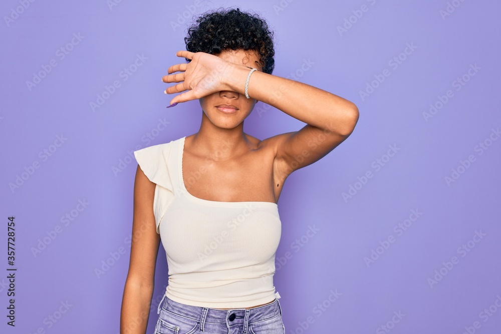 Young beautiful african american afro woman wearing casual t-shirt over purple background covering eyes with arm, looking serious and sad. Sightless, hiding and rejection concept Young beautiful african american afro woman wearing casual t-shirt over purple background covering eyes with arm, looking serious and sad. Sightless, hiding and rejection concept