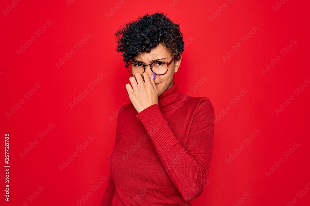 Young beautiful african american afro woman wearing turtleneck sweater and glasses smelling something stinky and disgusting, intolerable smell, holding breath with fingers on nose. Bad smell Young beautiful african american afro woman wearing turtleneck sweater and glasses smelling something stinky and disgusting, intolerable smell, holding breath with fingers on nose. Bad smell