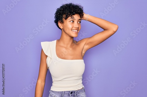 Young beautiful african american afro woman wearing casual t-shirt over purple background smiling confident touching hair with hand up gesture, posing attractive and fashionable