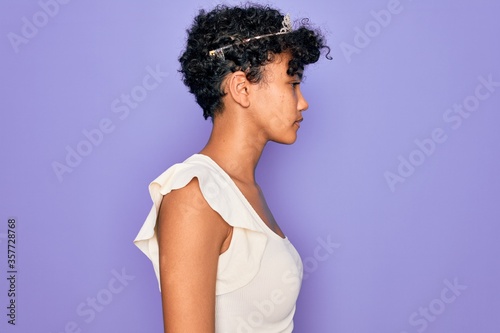 Young beautiful african american afro woman wearing tiara crown over purple background looking to side, relax profile pose with natural face with confident smile.
