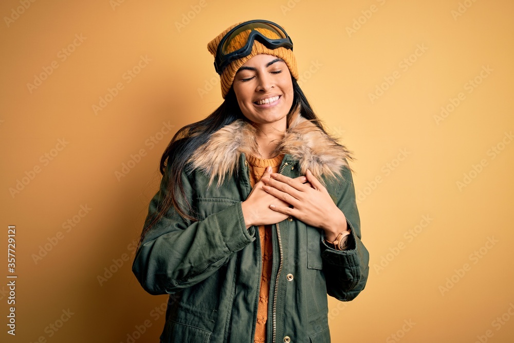 Young beautiful hispanic woman wearing ski glasses and coat for winter weather smiling with hands on chest with closed eyes and grateful gesture on face. Health concept. Young beautiful hispanic woman wearing ski glasses and coat for winter weather smiling with hands on chest with closed eyes and grateful gesture on face. Health concept.