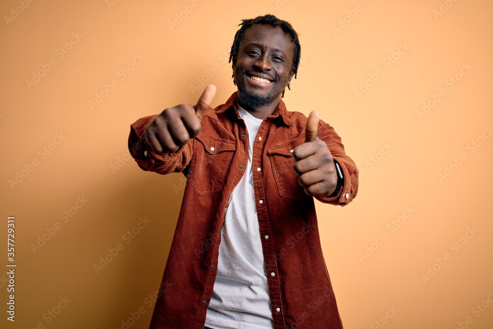 Young handsome african american man wearing casual jacket standing over yellow background approving doing positive gesture with hand, thumbs up smiling and happy for success. Winner gesture.