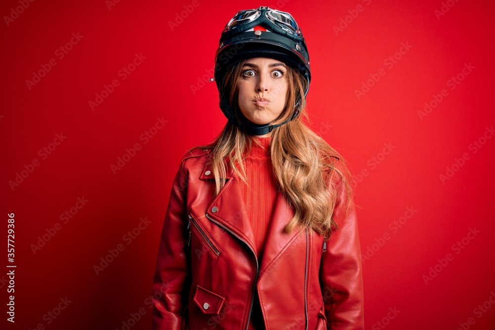 Young beautiful brunette motrocyclist woman wearing moto helmet over red background puffing cheeks with funny face. Mouth inflated with air, crazy expression. Young beautiful brunette motrocyclist woman wearing moto helmet over red background puffing cheeks with funny face. Mouth inflated with air, crazy expression.