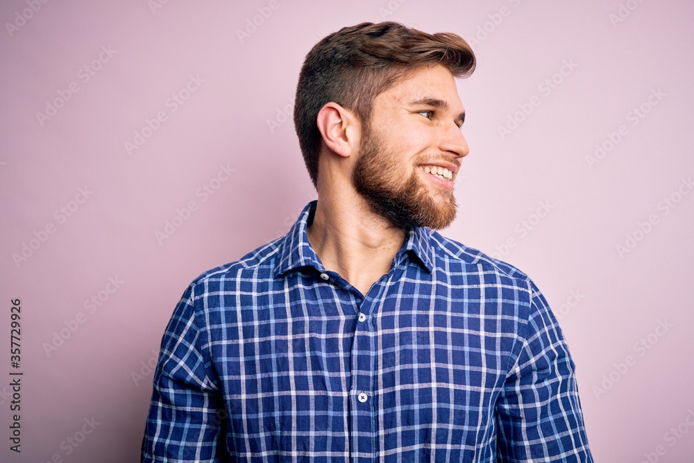 Young handsome blond man with beard and blue eyes wearing casual shirt standing looking away to side with smile on face, natural expression. Laughing confident. Young handsome blond man with beard and blue eyes wearing casual shirt standing looking away to side with smile on face, natural expression. Laughing confident.