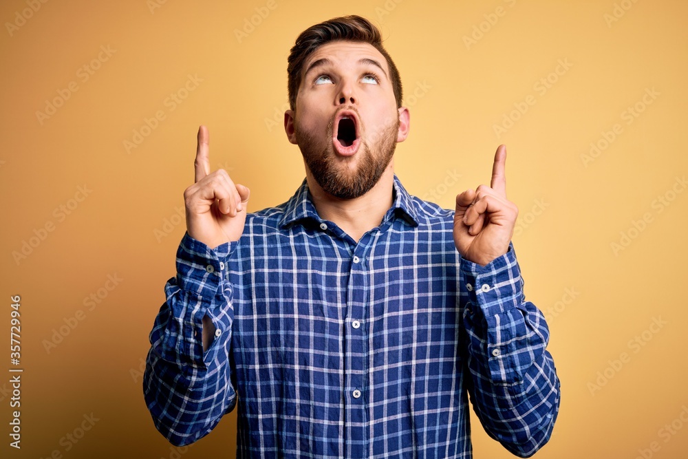 Young blond businessman with beard and blue eyes wearing shirt over yellow background amazed and surprised looking up and pointing with fingers and raised arms. Young blond businessman with beard and blue eyes wearing shirt over yellow background amazed and surprised looking up and pointing with fingers and raised arms.