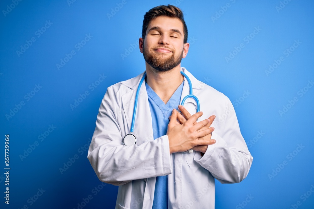 Young blond doctor man with beard and blue eyes wearing white coat and stethoscope smiling with hands on chest with closed eyes and grateful gesture on face. Health concept. Young blond doctor man with beard and blue eyes wearing white coat and stethoscope smiling with hands on chest with closed eyes and grateful gesture on face. Health concept.