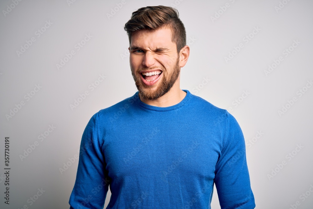 Young handsome blond man with beard and blue eyes wearing casual sweater winking looking at the camera with sexy expression, cheerful and happy face. Young handsome blond man with beard and blue eyes wearing casual sweater winking looking at the camera with sexy expression, cheerful and happy face.