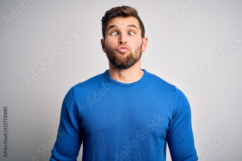 Young handsome blond man with beard and blue eyes wearing casual sweater making fish face with lips, crazy and comical gesture. Funny expression.