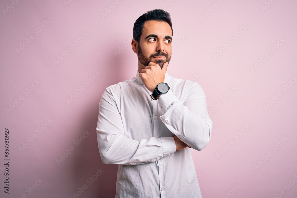 Young handsome man with beard wearing casual shirt standing over pink background with hand on chin thinking about question, pensive expression. Smiling with thoughtful face. Doubt concept. Young handsome man with beard wearing casual shirt standing over pink background with hand on chin thinking about question, pensive expression. Smiling with thoughtful face. Doubt concept.