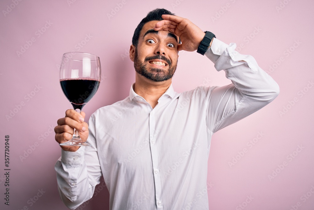 Young handsome man with beard drinking glass of red wine over isolated pink background stressed with hand on head, shocked with shame and surprise face, angry and frustrated. Fear and upset Young handsome man with beard drinking glass of red wine over isolated pink background stressed with hand on head, shocked with shame and surprise face, angry and frustrated. Fear and upset