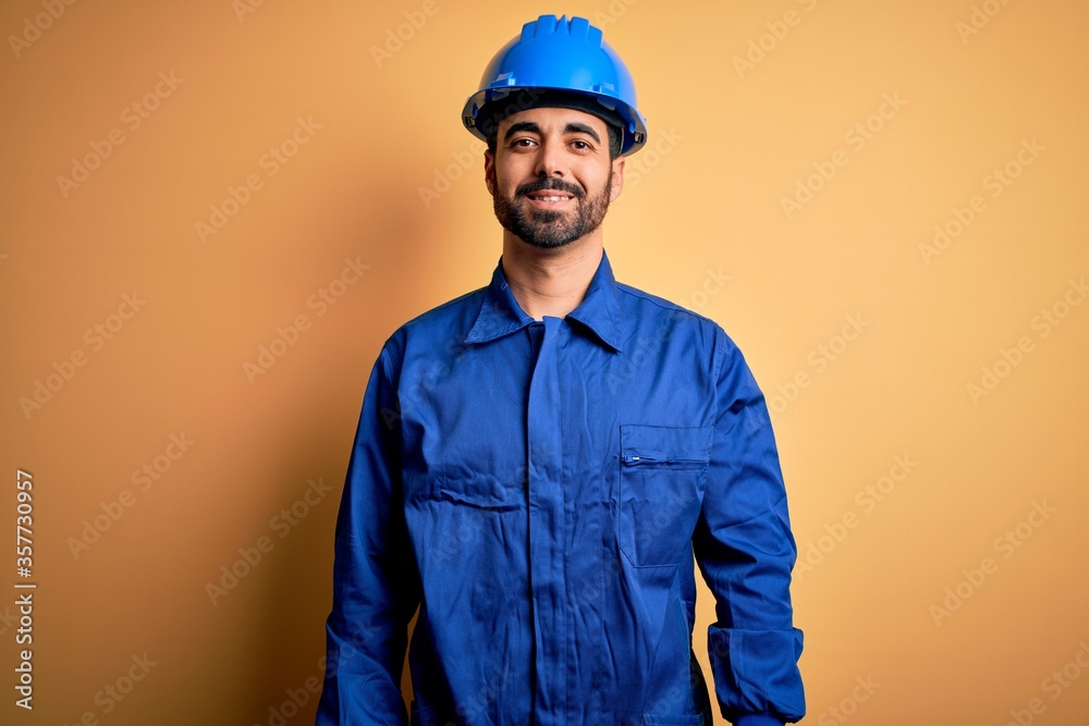 Mechanic man with beard wearing blue uniform and safety helmet over yellow background with a happy and cool smile on face. Lucky person.