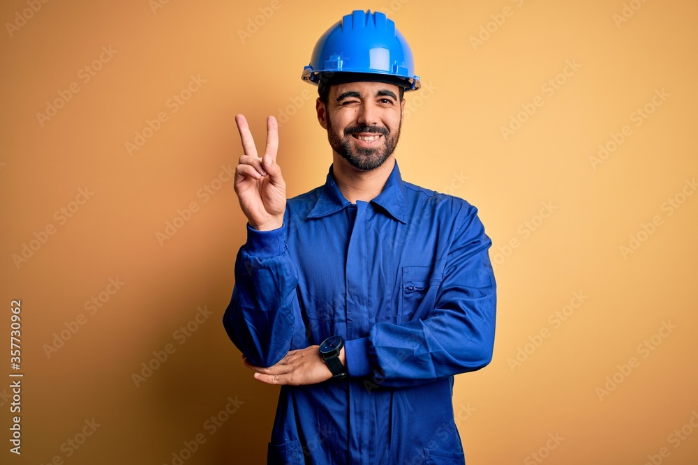 Mechanic man with beard wearing blue uniform and safety helmet over yellow background smiling with happy face winking at the camera doing victory sign with fingers. Number two.