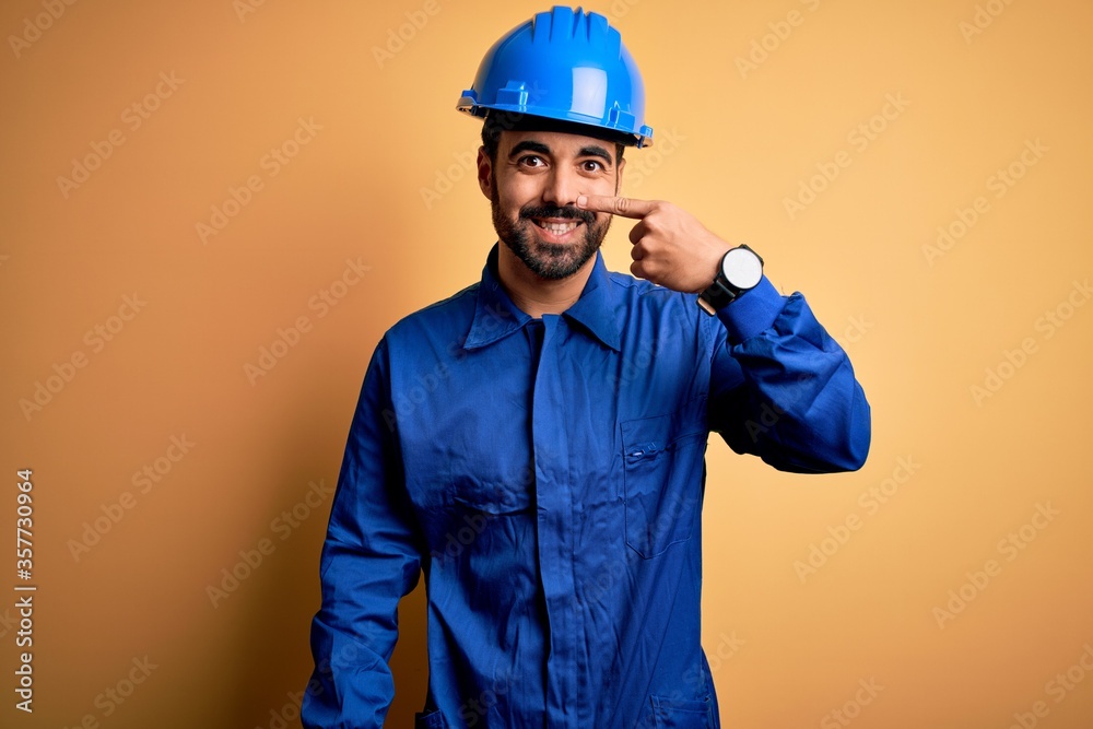 Mechanic man with beard wearing blue uniform and safety helmet over yellow background Pointing with hand finger to face and nose, smiling cheerful. Beauty concept
