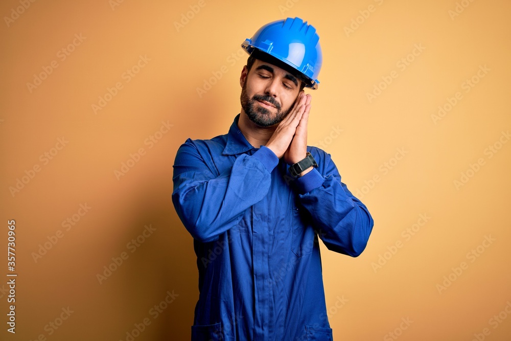 Mechanic man with beard wearing blue uniform and safety helmet over yellow background sleeping tired dreaming and posing with hands together while smiling with closed eyes.