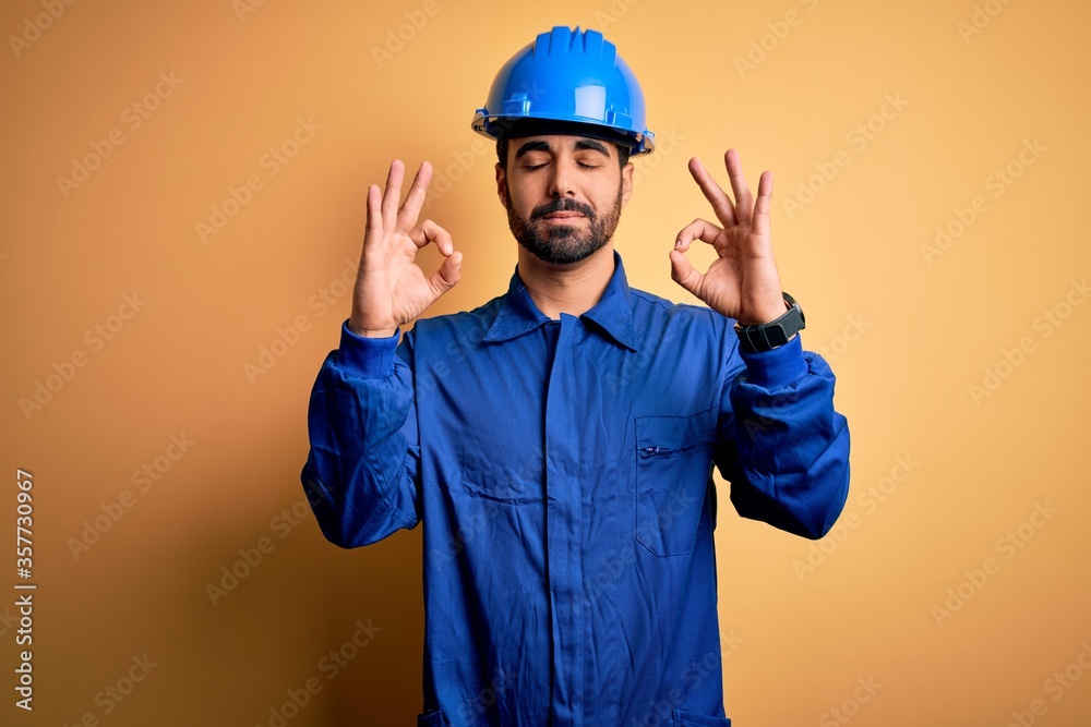 Mechanic man with beard wearing blue uniform and safety helmet over yellow background relaxed and smiling with eyes closed doing meditation gesture with fingers. Yoga concept.