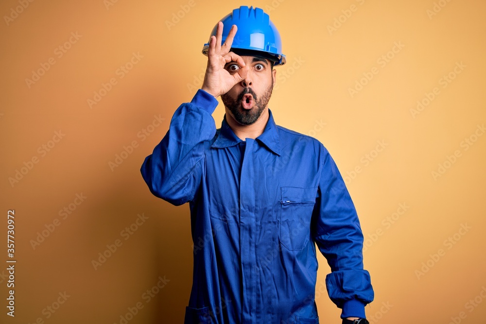 Mechanic man with beard wearing blue uniform and safety helmet over yellow background doing ok gesture shocked with surprised face, eye looking through fingers. Unbelieving expression. Mechanic man with beard wearing blue uniform and safety helmet over yellow background doing ok gesture shocked with surprised face, eye looking through fingers. Unbelieving expression.