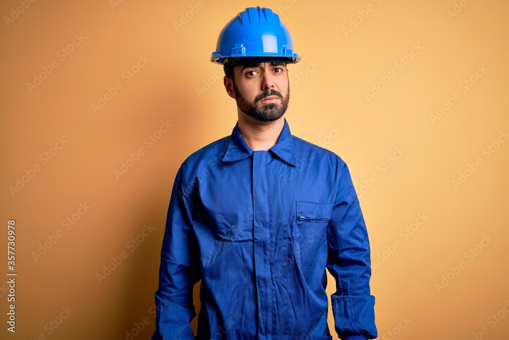 Mechanic man with beard wearing blue uniform and safety helmet over yellow background skeptic and nervous, frowning upset because of problem. Negative person.