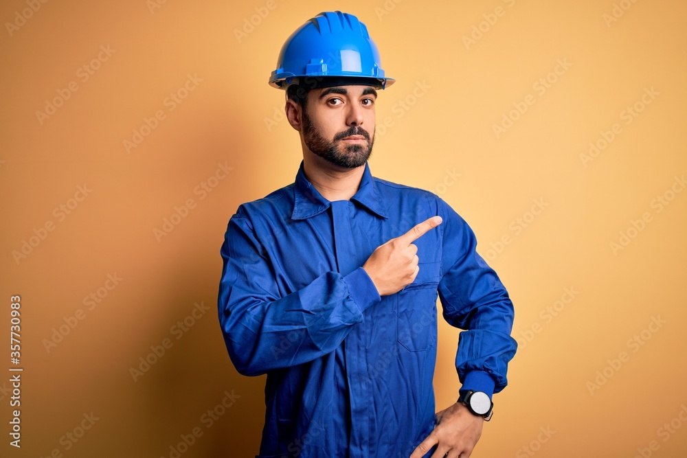 Mechanic man with beard wearing blue uniform and safety helmet over yellow background Pointing with hand finger to the side showing advertisement, serious and calm face
