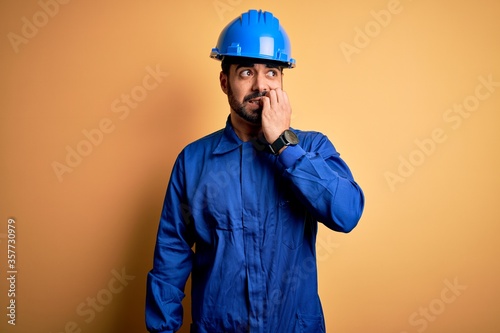 Mechanic man with beard wearing blue uniform and safety helmet over yellow background looking stressed and nervous with hands on mouth biting nails. Anxiety problem.