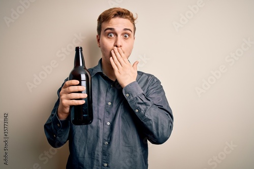 Young handsome redhead man drinking bottle of beer over isolated white background covering mouth with hand, shocked and afraid for mistake. Surprised expression