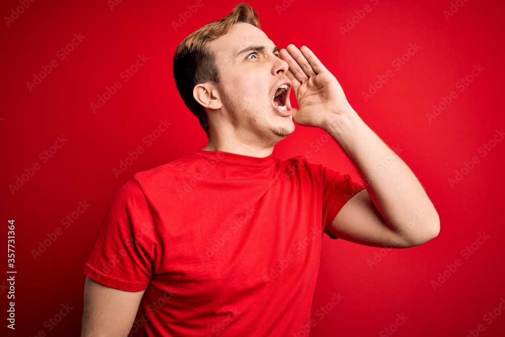 Young handsome redhead man wearing casual t-shirt over isolated red background shouting and screaming loud to side with hand on mouth. Communication concept.