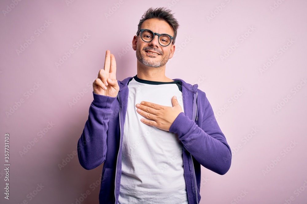 Young handsome man wearing purple sweatshirt and glasses standing over pink background smiling swearing with hand on chest and fingers up, making a loyalty promise oath