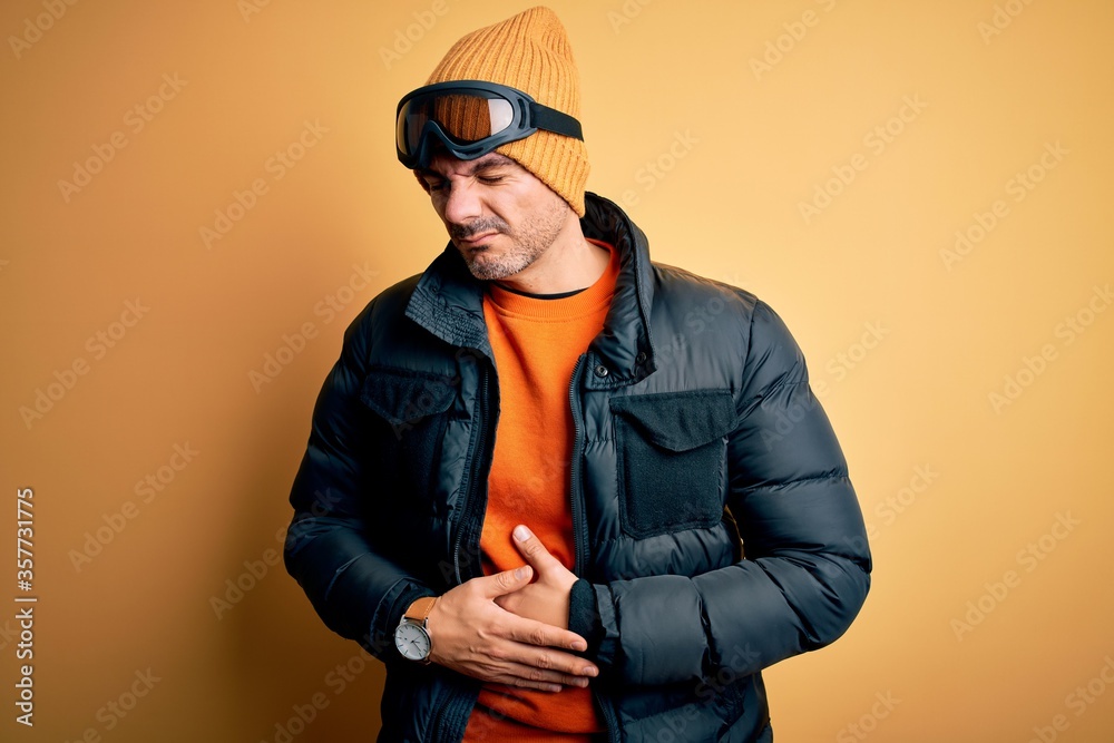 Young handsome skier man skiing wearing snow sportswear using ski goggles with hand on stomach because indigestion, painful illness feeling unwell. Ache concept. Young handsome skier man skiing wearing snow sportswear using ski goggles with hand on stomach because indigestion, painful illness feeling unwell. Ache concept.
